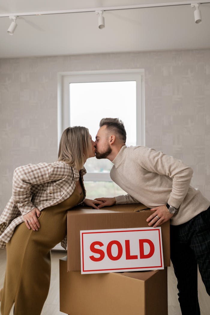 About Joyful couple kissing in their new home with 'sold' sign and boxes.